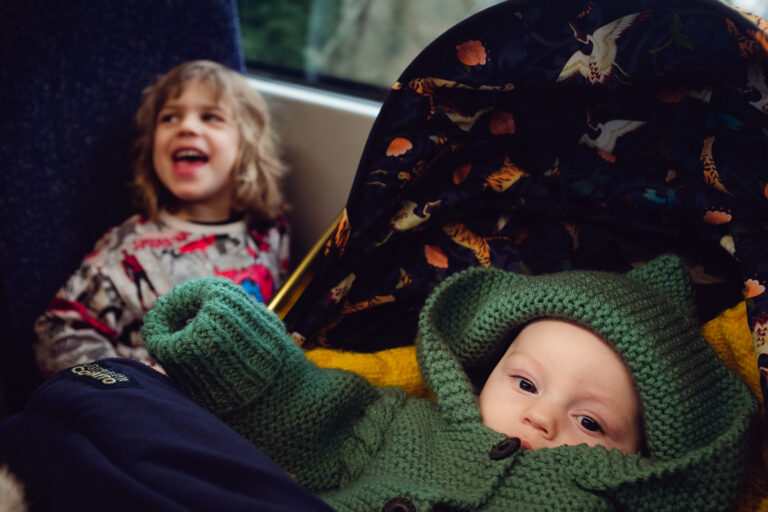 A baby in a knitted green onesie lying in a pram and a young boy behind laughing on a train to London