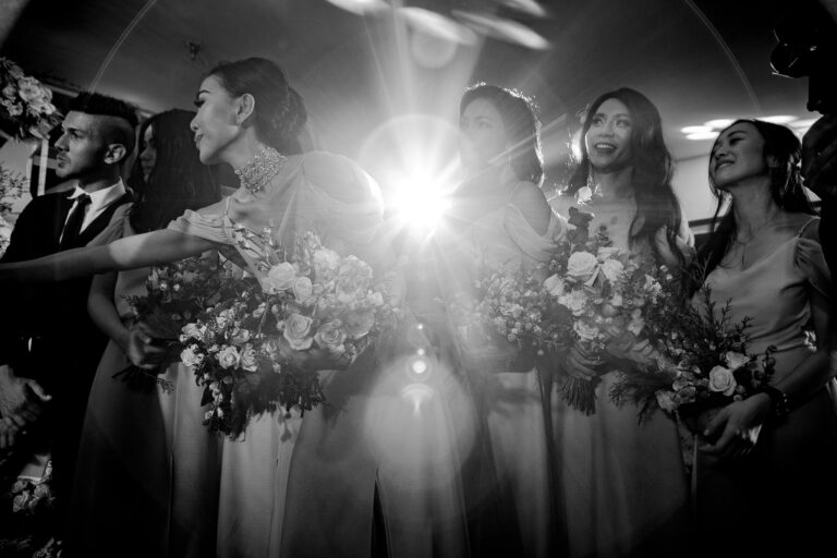 Bridesmaids holding big bouquets with a bright spotlight behind them at a wedding ceremony.