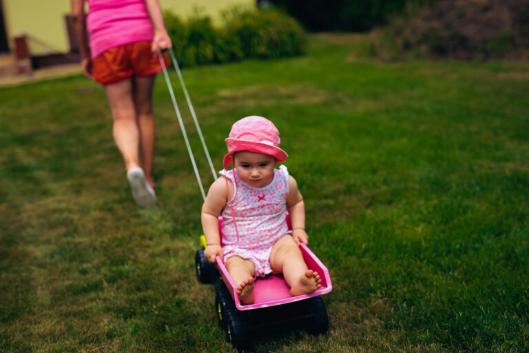 A toddler in a pink onesie being pulled on a pink truck by her grandma through the grass