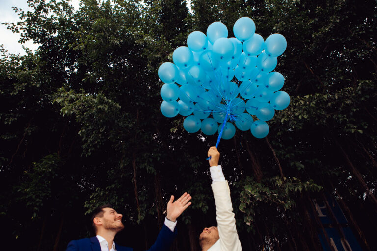 LGBTQ+ couple holding blue balloons into the air just after their wedding ceremony.
