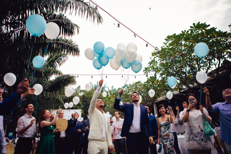 LGBTQ+ couple and guests holding lots of blue and white balloons at a wedding ceremony.