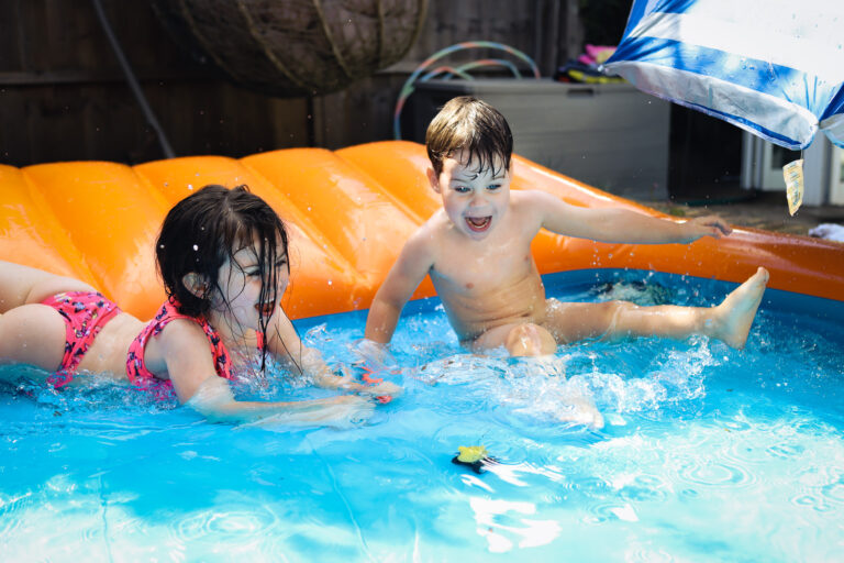 A young boy and girl laughing and sliding into a paddling pool on a summers day in London, UK
