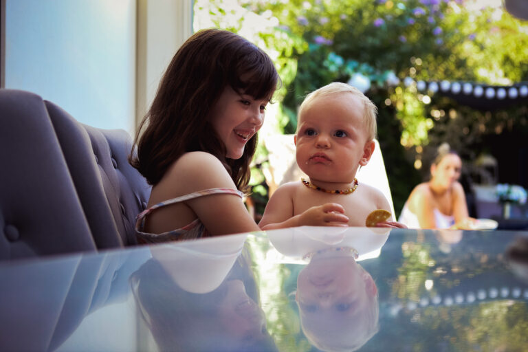 A young girl laughing at a table holding a baby who's eating a biscuit on a warm summers day