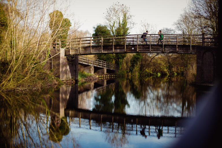 family portrait of mum dad and toddler playing up on a bridge with the reflection in the river below