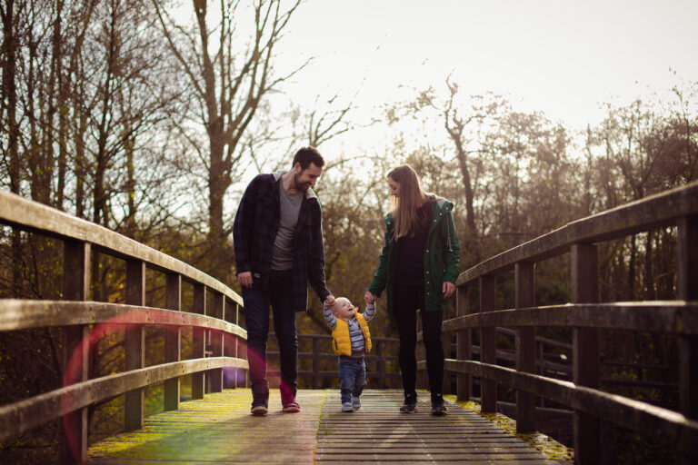 family portrait of mum and dad holding toddlers hands as they walk across a wooden bridge