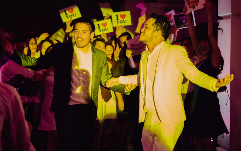 LGBTQ+ dancing at their wedding reception surrounded by guests holding I love you placards.