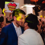 LGBTQ+ couple kissing surrounded by guests holding just married signs at their wedding reception.