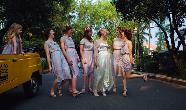 Bride and bridesmaids laughing together in a row whilst a girl looks at them from the car.