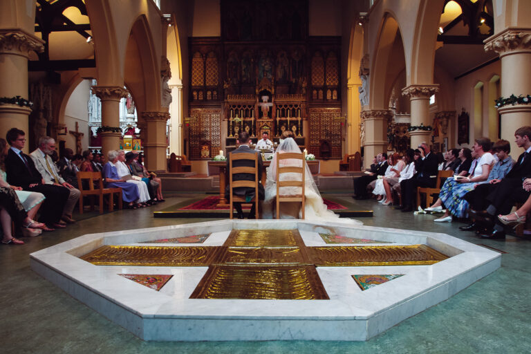 Wide angle shot of the bride and groom sitting on chairs listening to the priest at their church wedding ceremony.