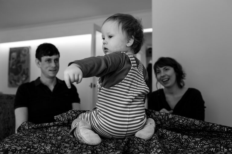 A toddler sitting on a table with mum and dad smiling in the background during a family photo session