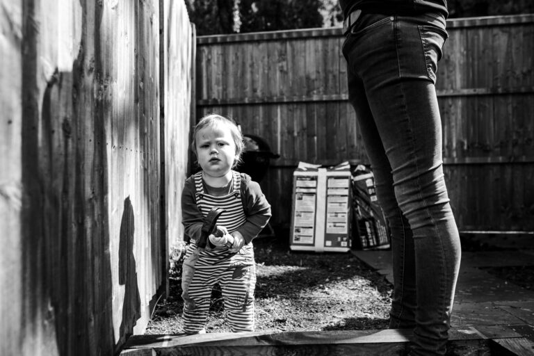 A toddler lifting a heavy hammer in a garden with his dad next to him during a family photo session