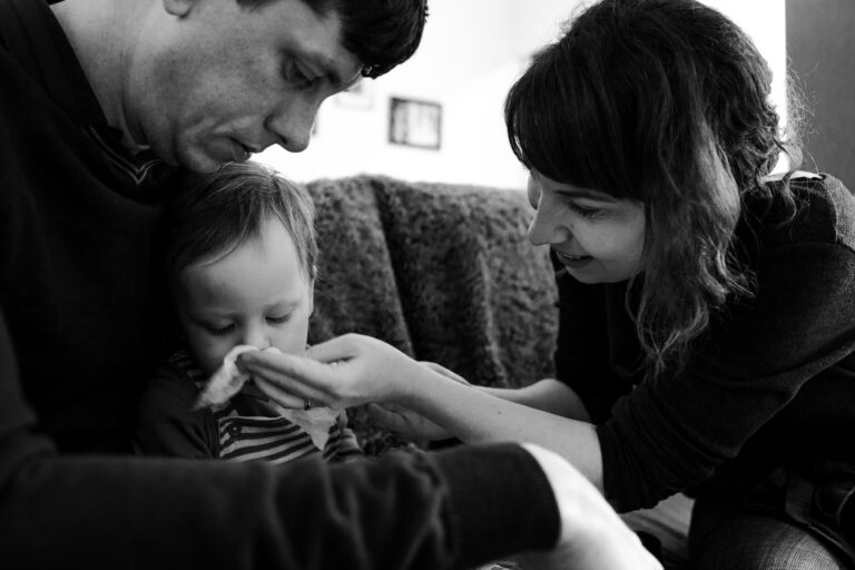 Mum and dad sit and wipe toddlers nose during a family photo session