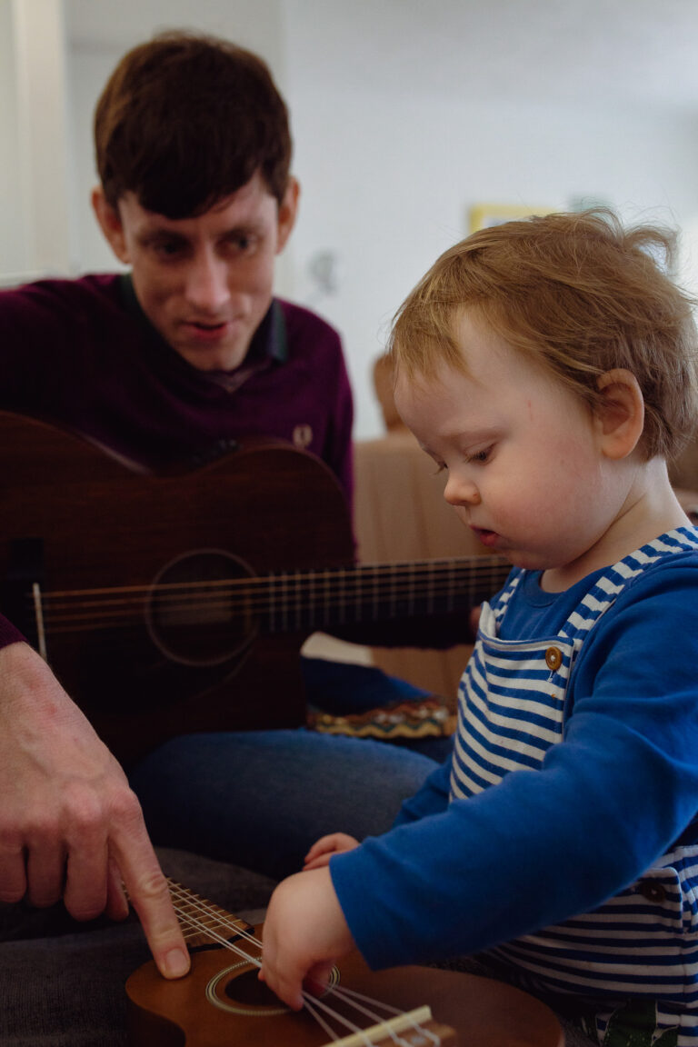 Dad holding a guitar and pointing to a ukelele teaching a toddler how to play during a family photo session