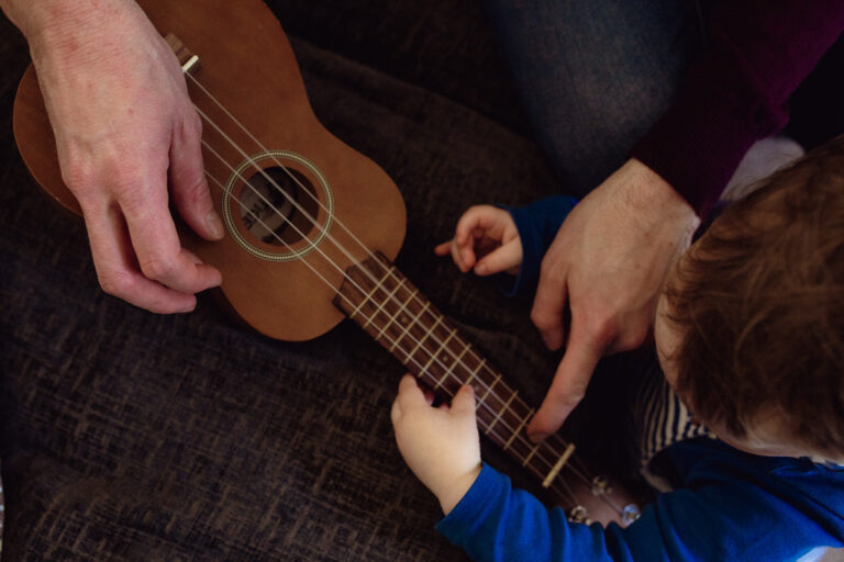 Dad's hands pointing to a ukulele and teaching his toddler about music during a family photo session