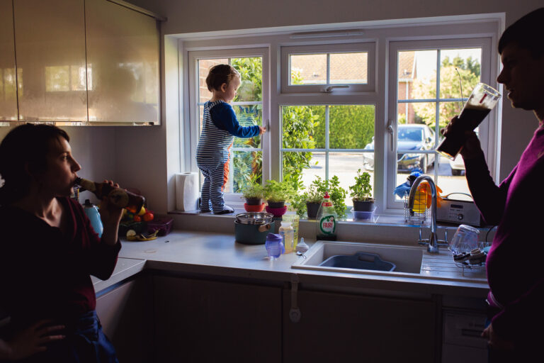 Mum and dad drinking beer in the kitchen with a toddler standing on a windowsill during a family photo session