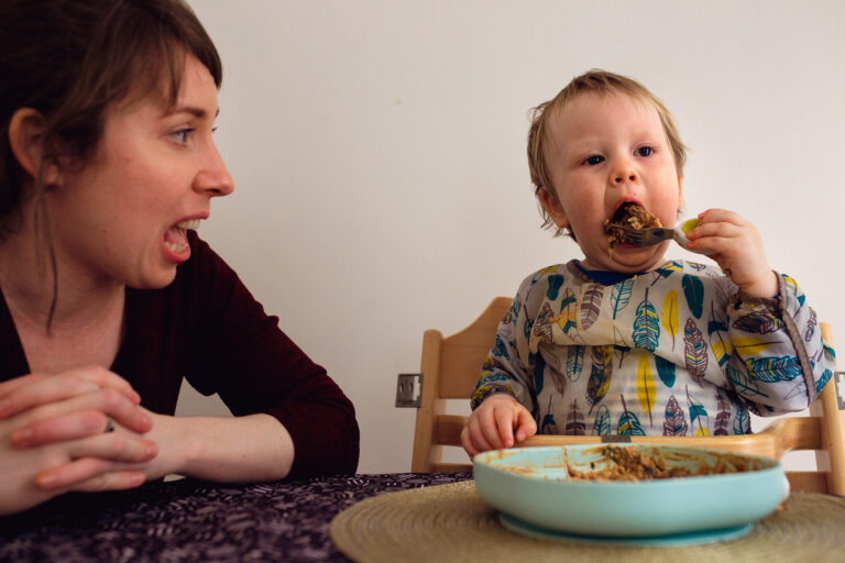 Mum reacting to toddler feeding himself at the dinner table during a family photo session
