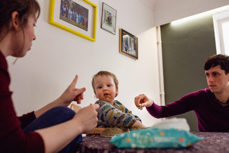 Messy toddler covered in food sitting at the family dinner table with mum and dad during a family photo session