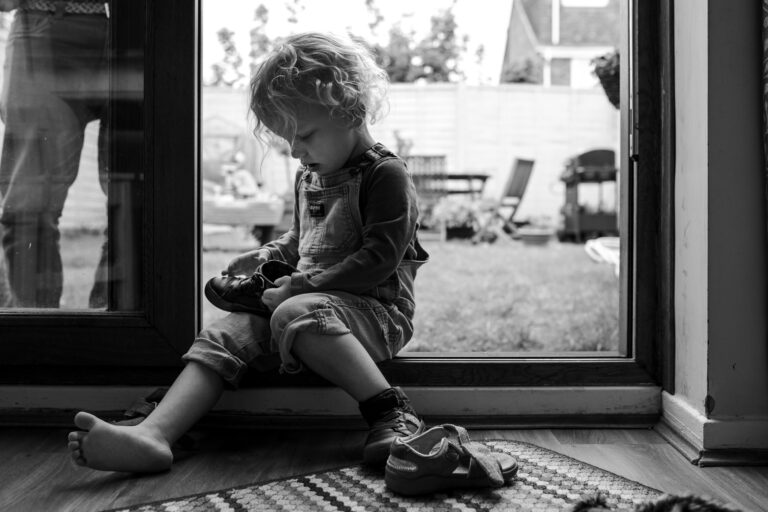 A young boy sits by a sliding door to the garden and puts on his shoes during a family photo session.