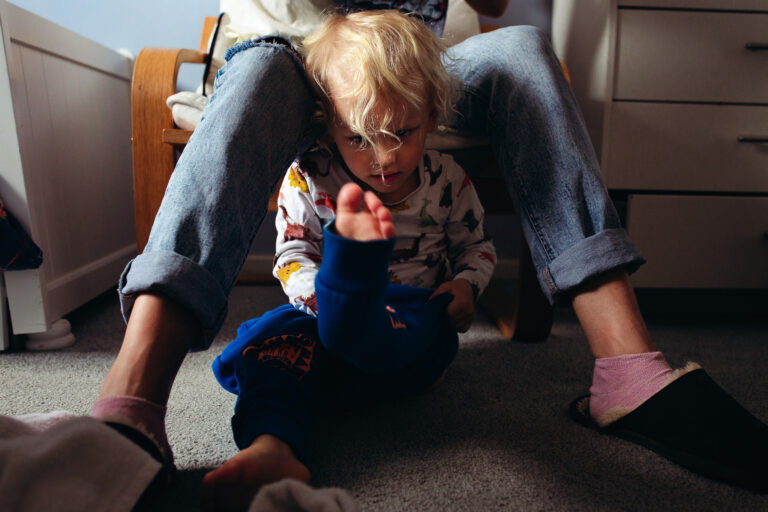 A young boy sitting in between his mums legs putting on his trousers during a family photo session