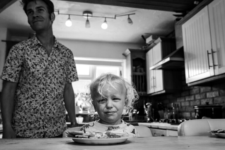 A young boy cries whilst eating at the kitchen counter with dad smiling behind him during a family photo session