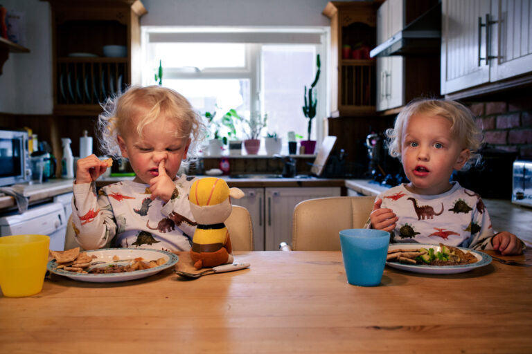 Two young boy twins eating dinner with one of them picking their nose during a family photo session