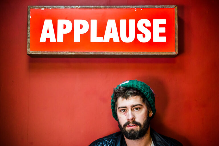 young man with a beard and green beanie standing under a large applause sign