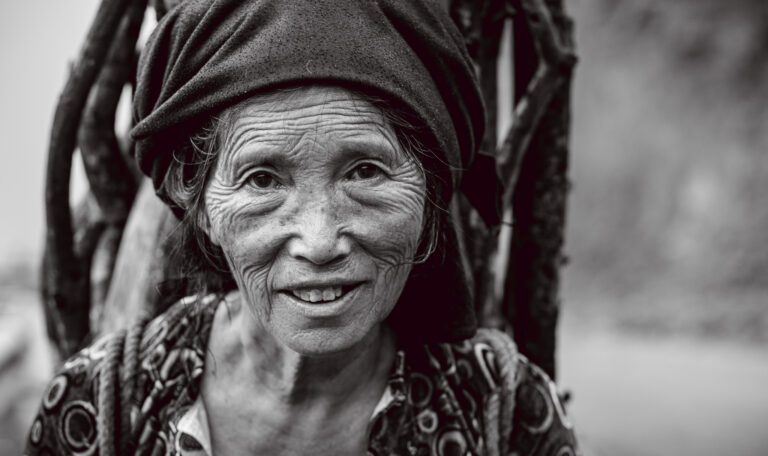 An elderly Vietnamese woman carrying a bag of sticks on her back poses for a portrait on the Ha Giang pass