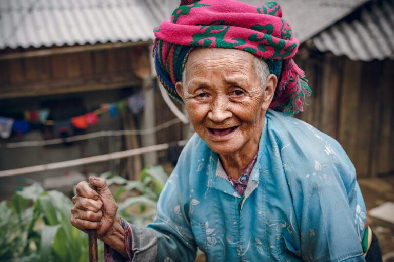 An elderly woman with a walking stick wearing a pink turban poses for a portrait on the Ha Giang pass