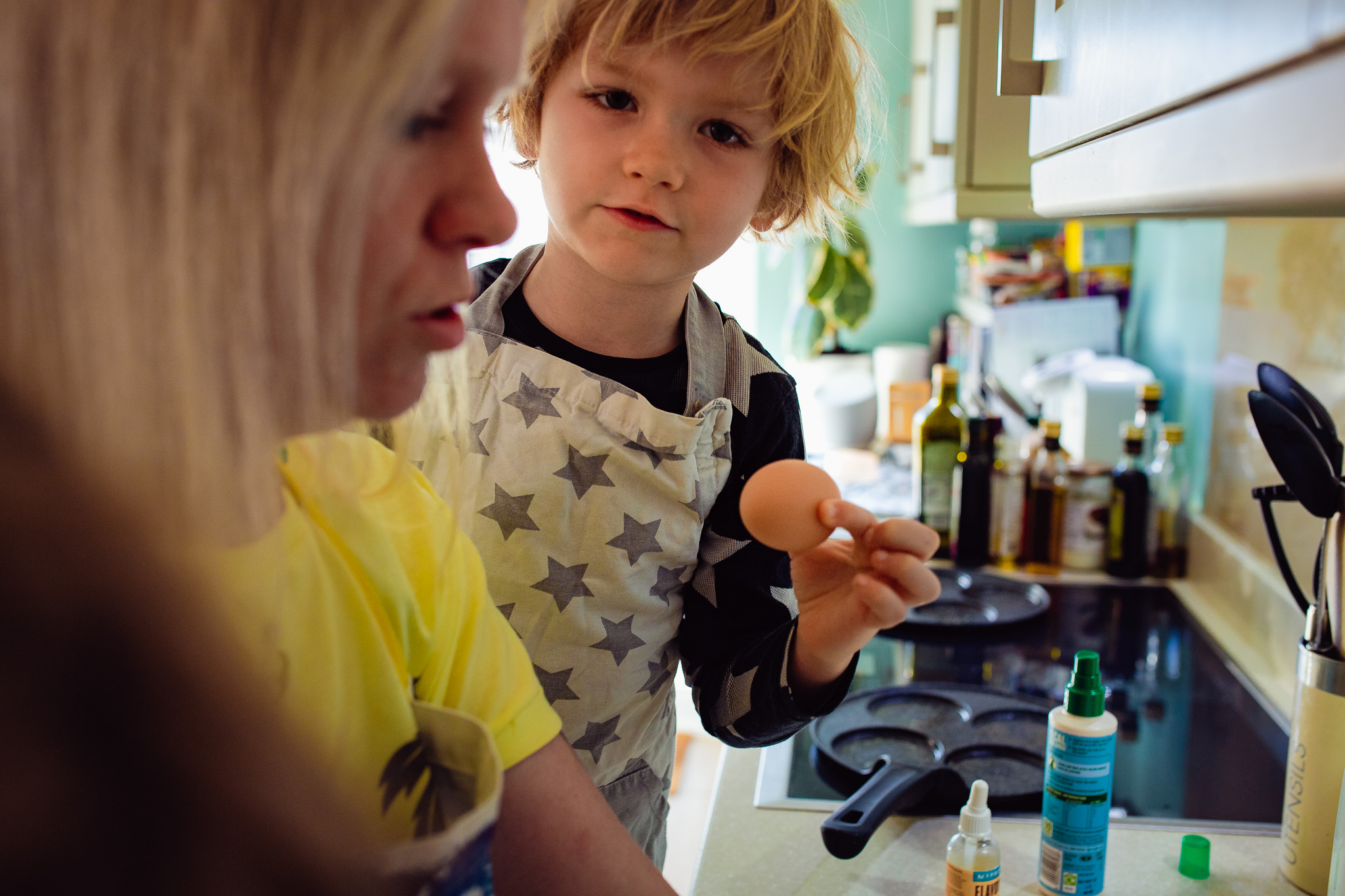 Young boy wearing an apron, holding an egg, helping his mom cook pancakes during a family photo session.