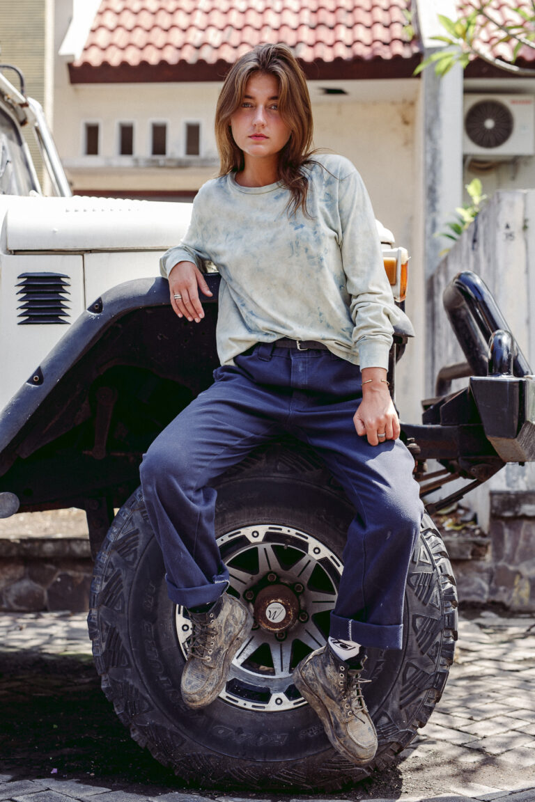 A young woman poses for a fashion portrait wearing a tie-dye jumper sitting on a wheel of a jeep