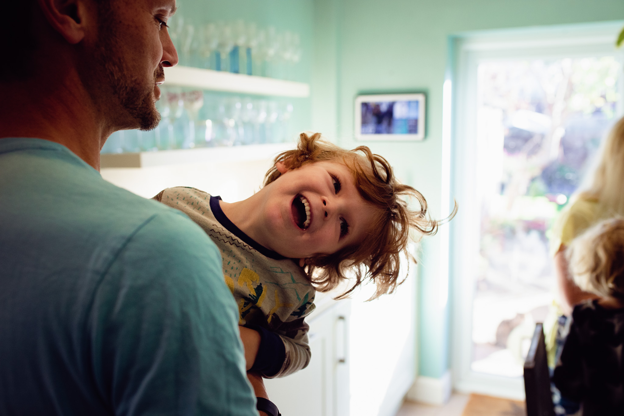 Dad and young son are laughing and playing in a turquoise kitchen during a family photo session.