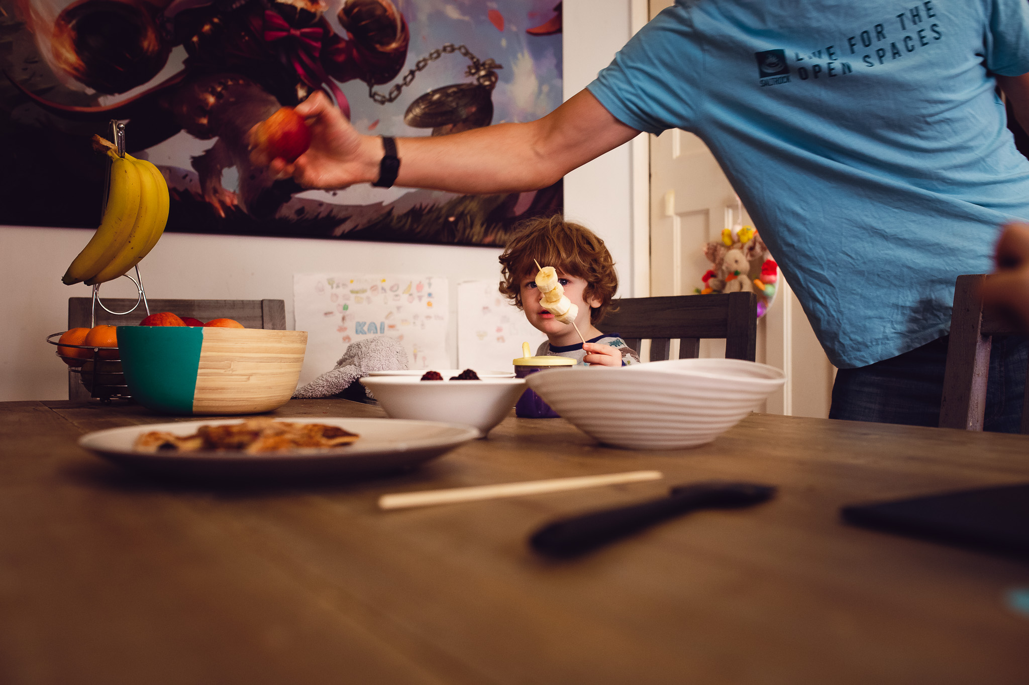 Leo sitting at a table with bananas on a skewer and dad reaching for an apple during a family photo session.