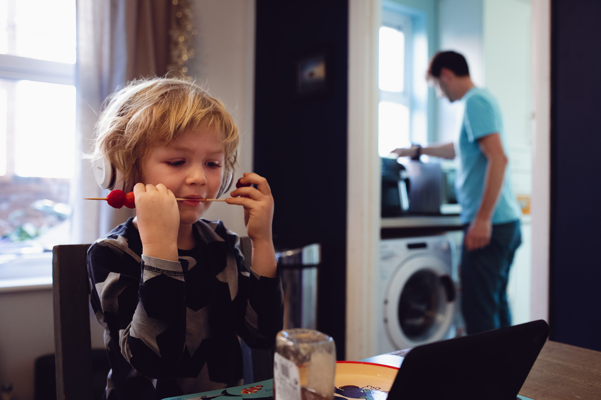 Young blonde boy wearing headphones, watching iPad, and eating fruit skewers during a family photo session.