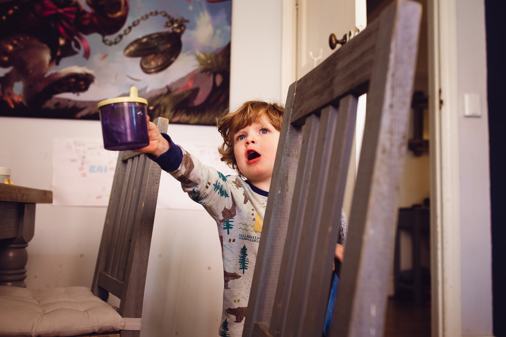 Young boy reaching up with a Tommee Tippee cup between two wooden chairs during a family photo session.