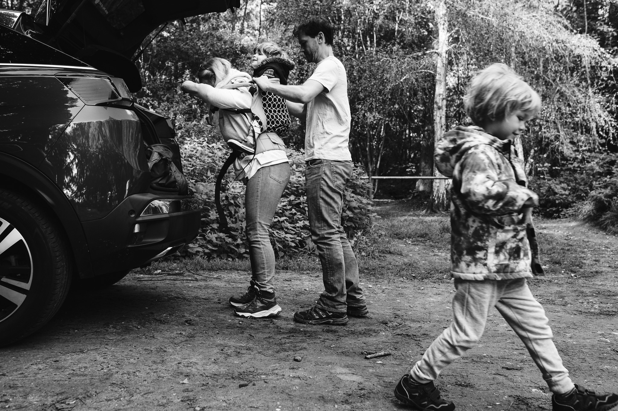 Dad is helping Mum put their son into a carrier on her back next to the trunk of the car during a family photo session.