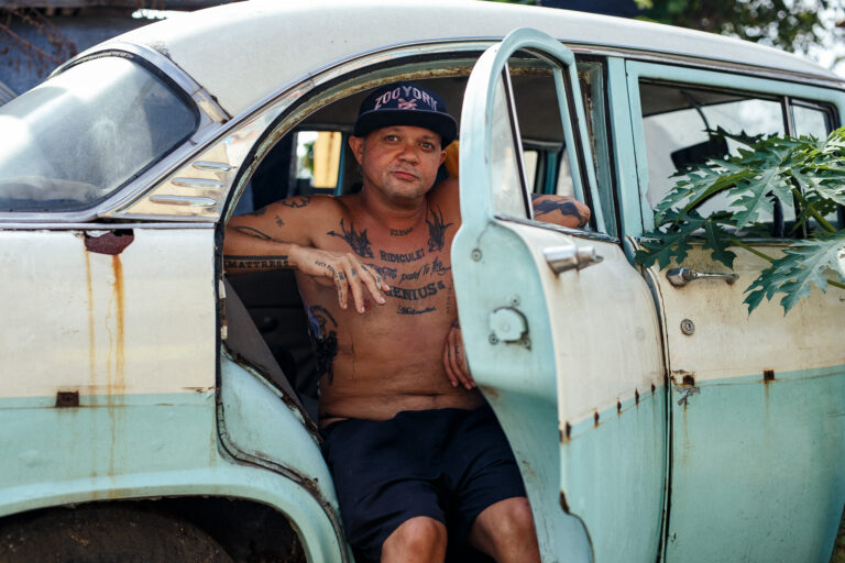 A tattooed topless man poses for a portrait sitting and smoking out of the back seat of an old car