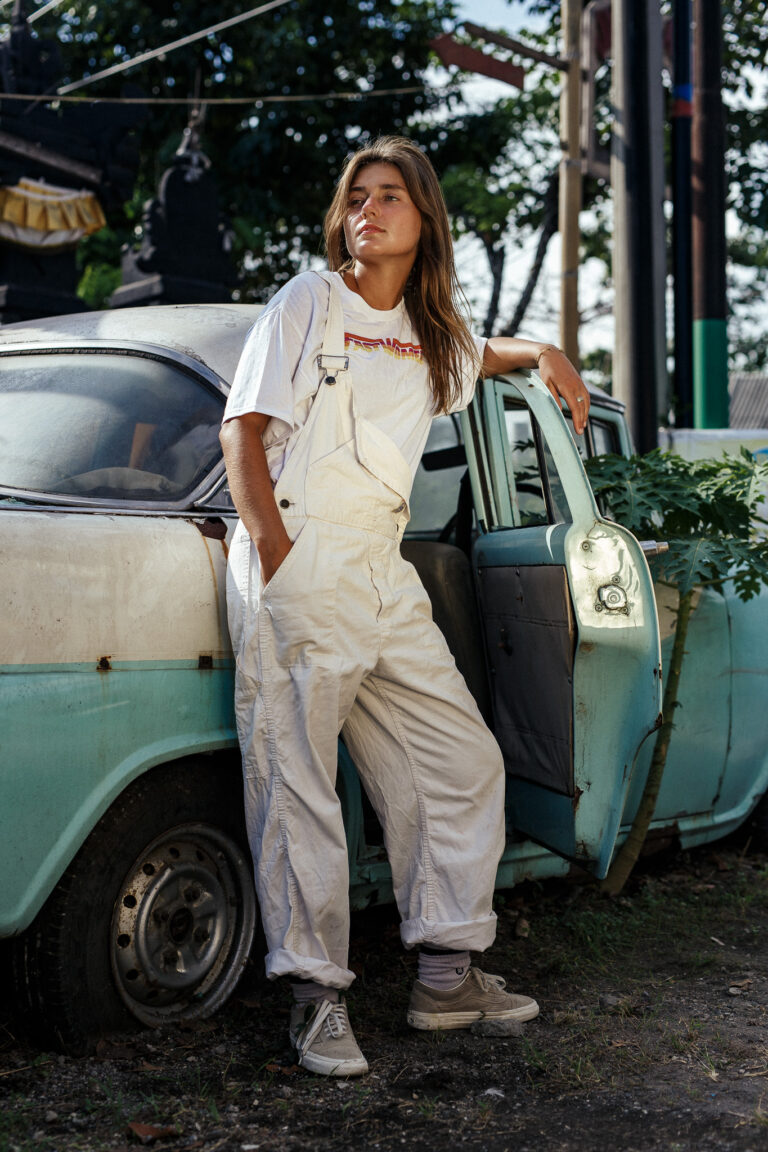 A young woman poses for a fashion portrait wearing white dungarees standing next to an old car