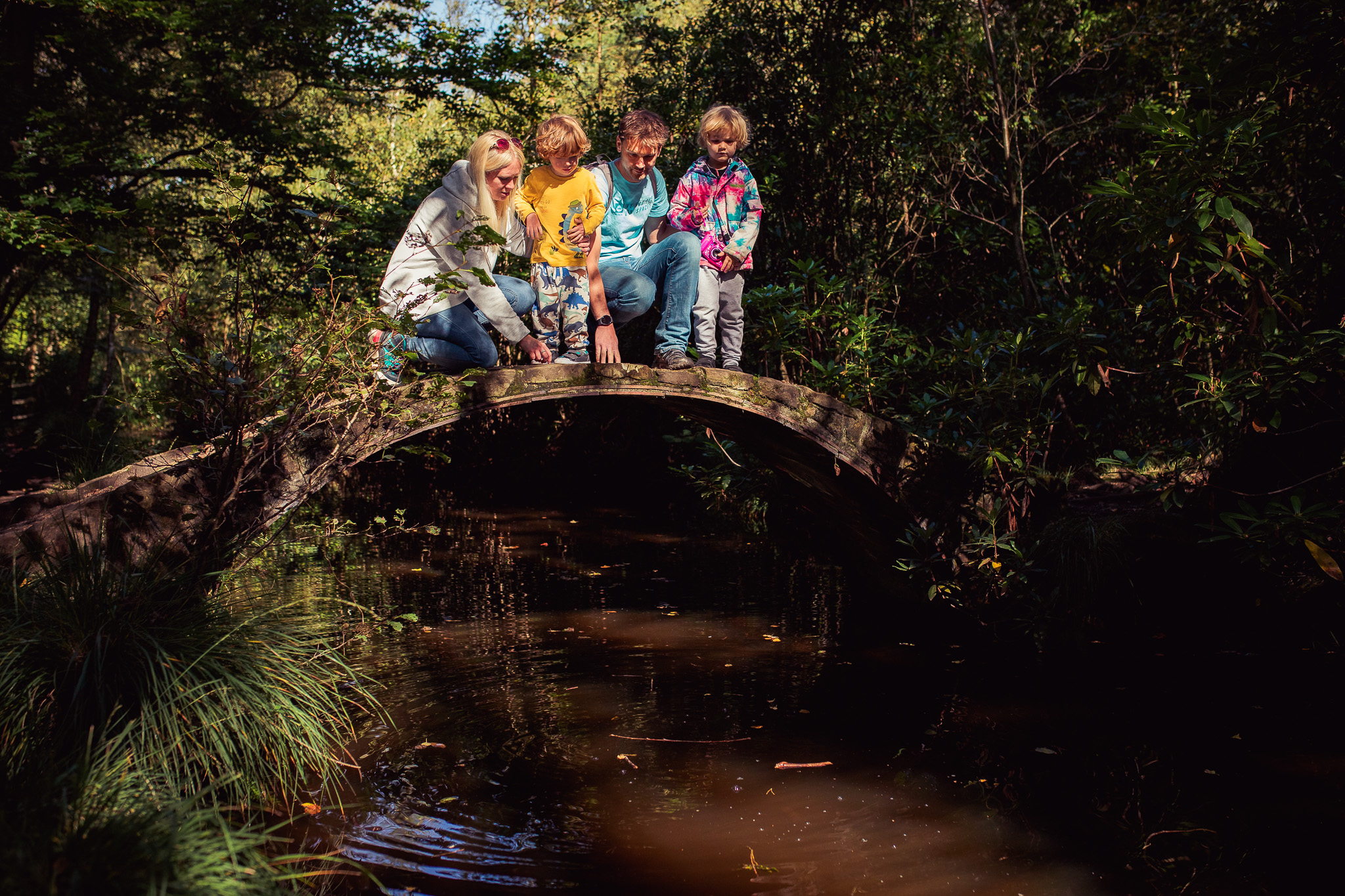 Mum, dad, and two sons are standing on a small stone bridge, looking over into the water during a family photo session.