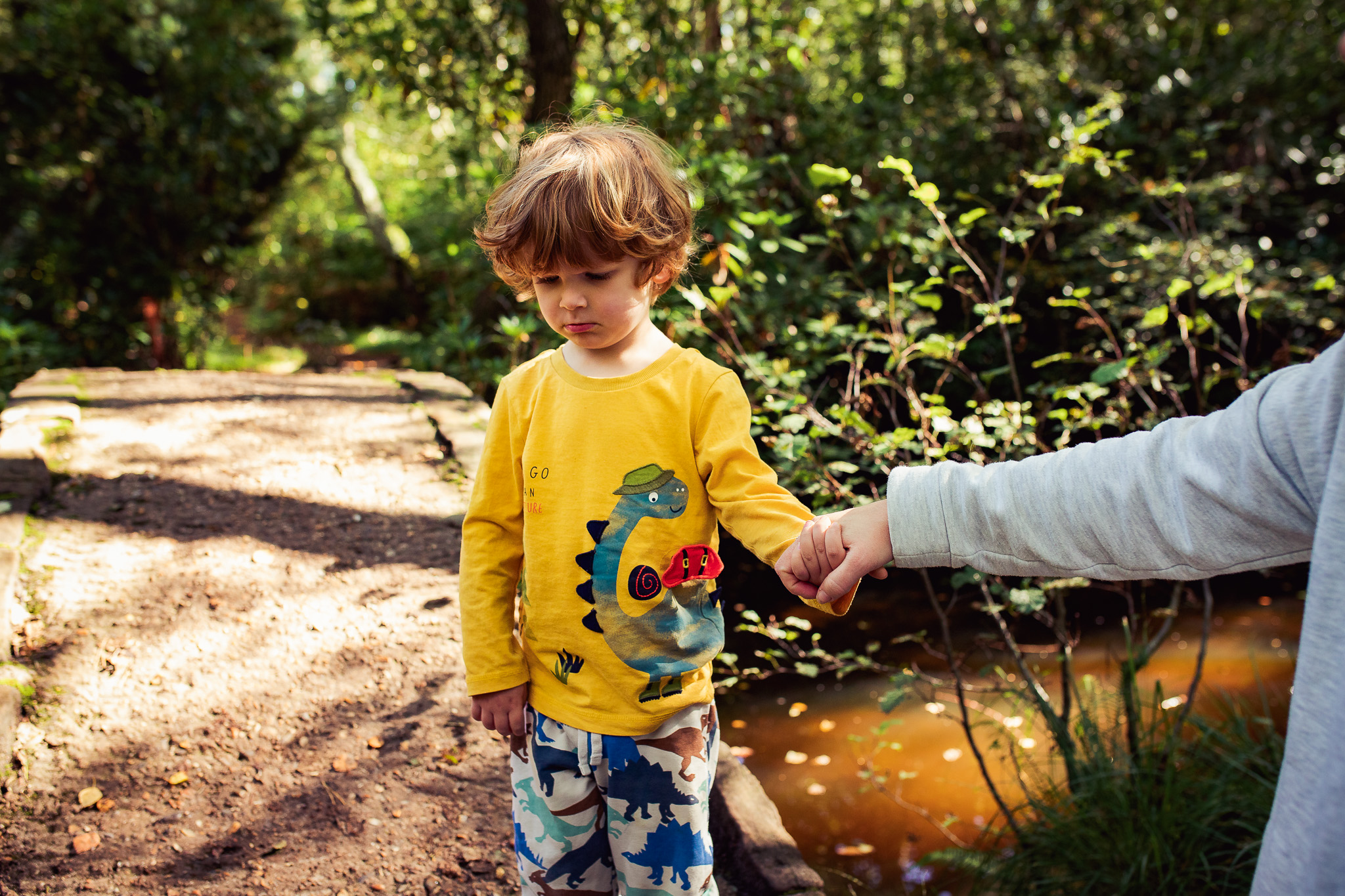 Leo stands next to a small stone bridge looking sad whilst holding mums hand during a family photo session.