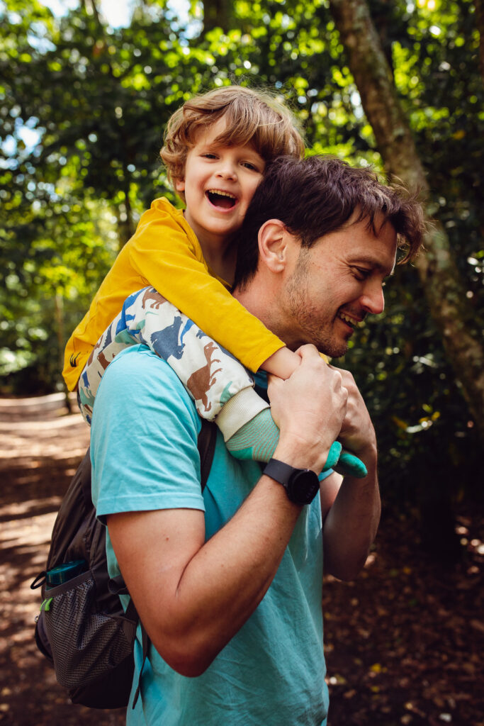 Dad carrying his son in a forest on his shoulders whilst laughing during a family photo session.