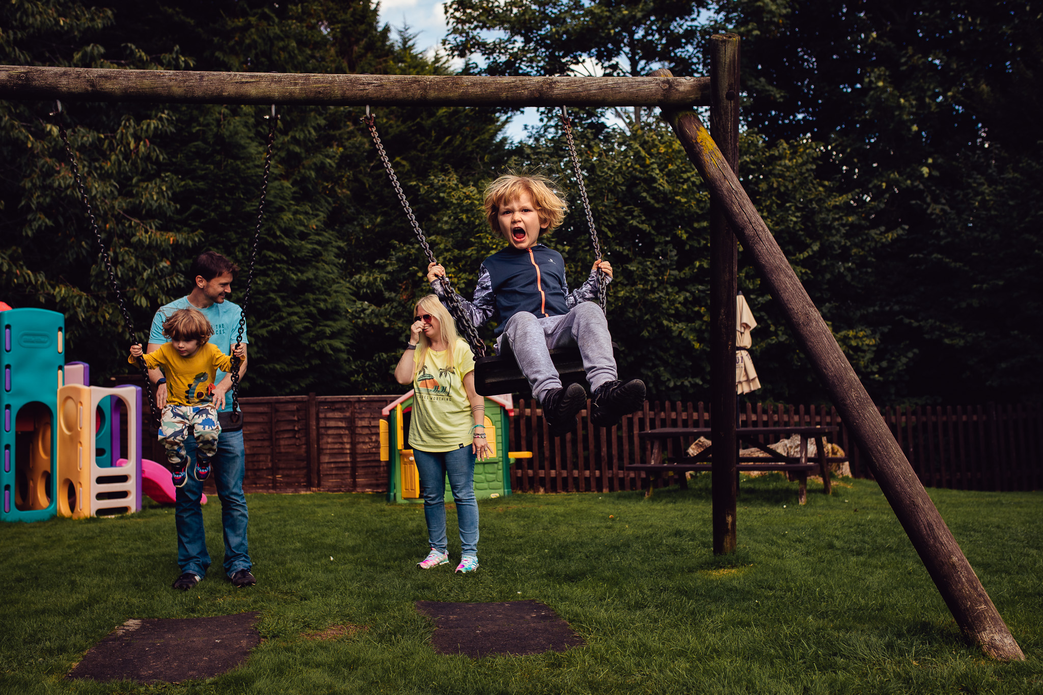 Mum and dad with their two sons on swings whilst one of the boys shouts during a family photo session.