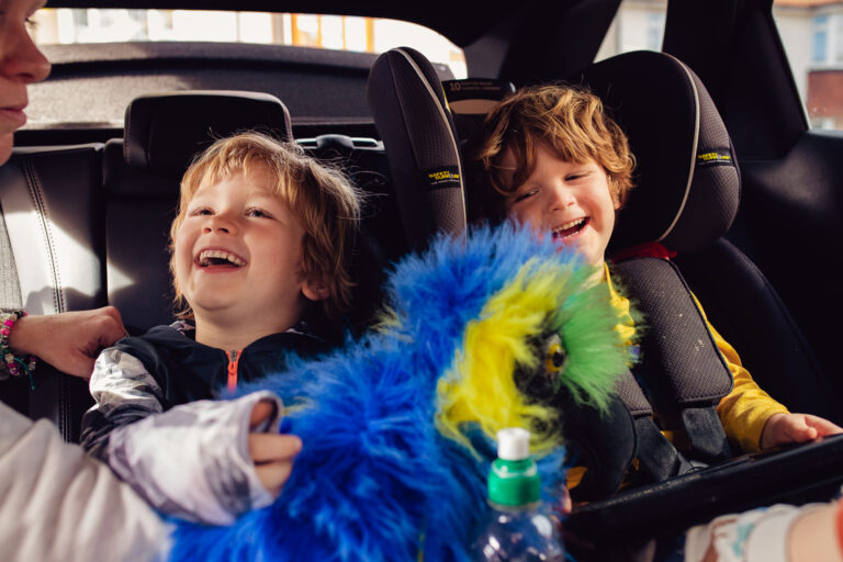 Kai and Leo laughing in the backseat of a car playing with their mum and a blue puppet.