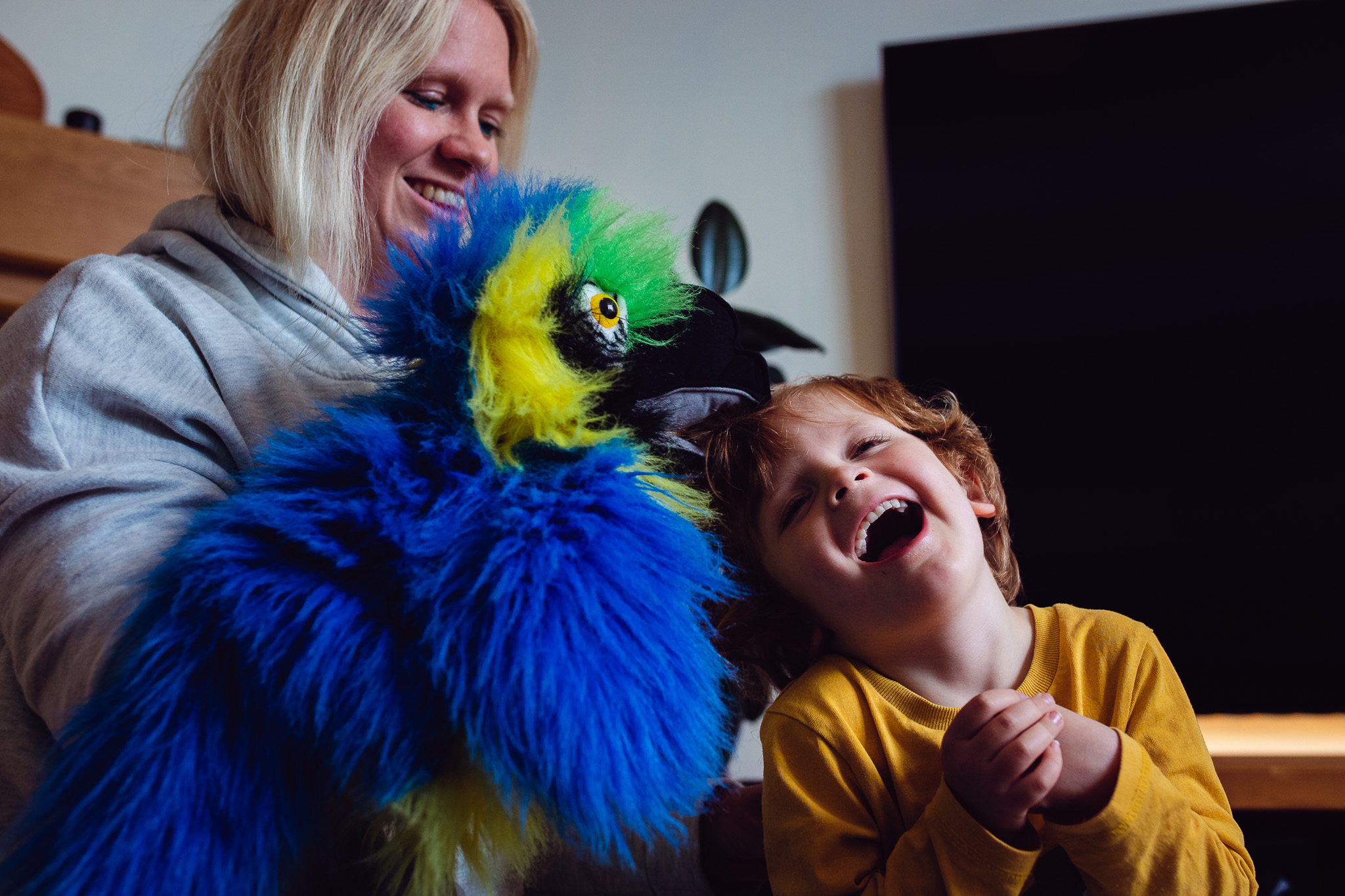 Mum holding a blue parrot hand puppet playing with her laughing son during a family photo session.