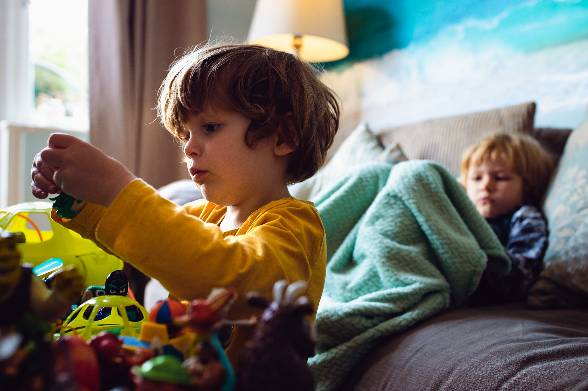 Leo playing with toys whilst Kai sits on sofa with his blanket during a family photo session.