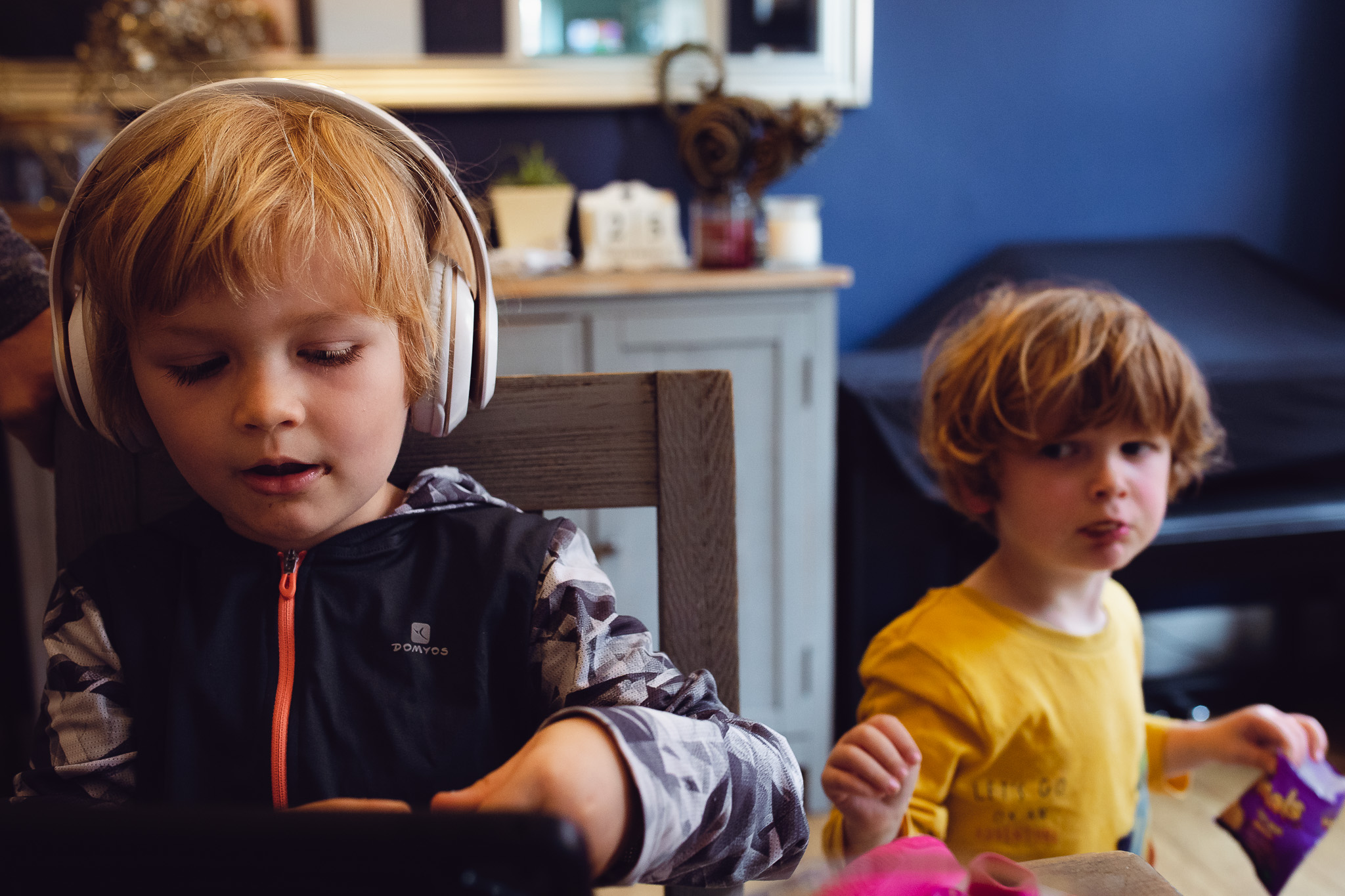 Kai sitting at a table wearing headphones while Leo walks behind him eating biscuits during a family photo session.