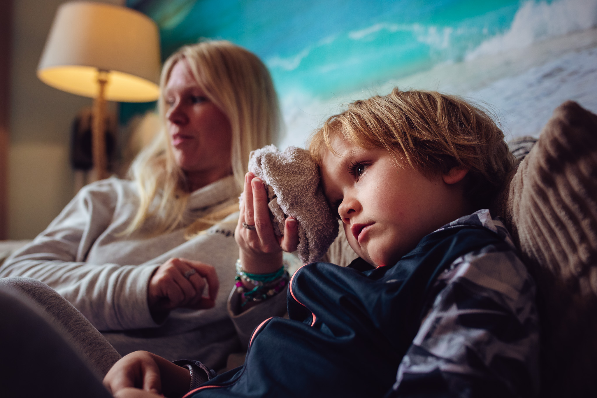 mum holding an iced flannel to a sad young boys head whilst sitting on a sofa during a family photo session.