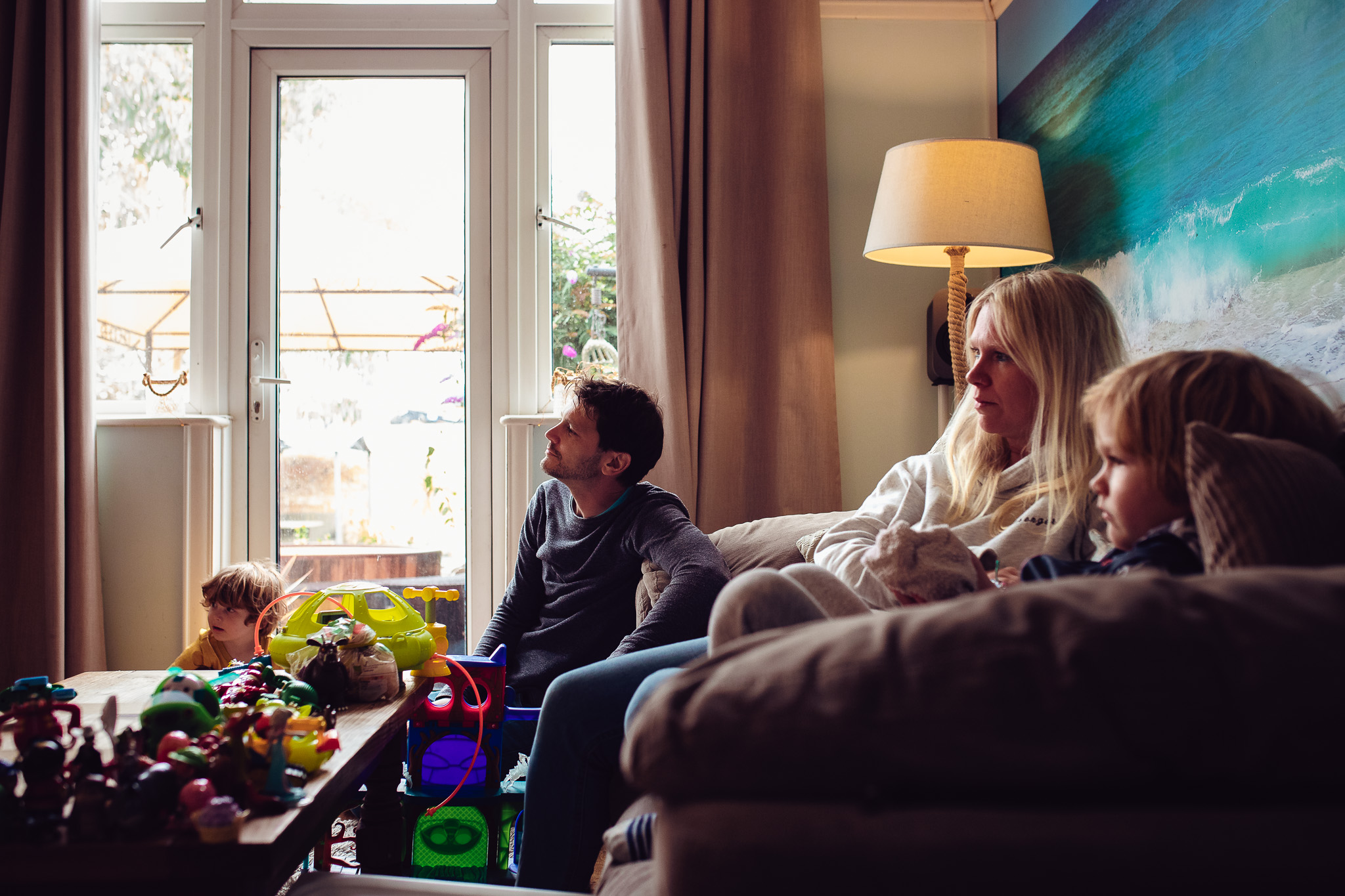 Mum, dad and 2 young boys watching tv in their living room during a family photo session.