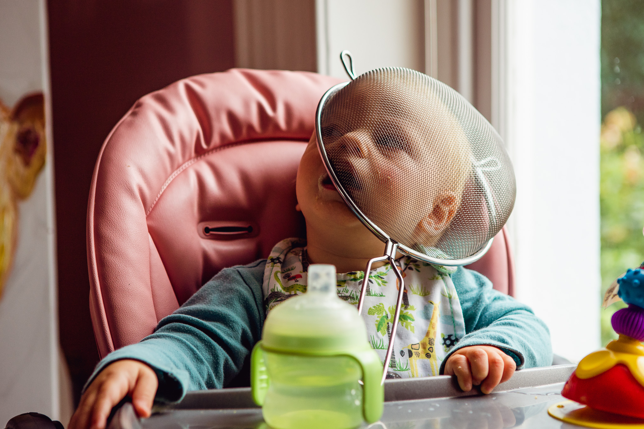 portrait of a baby sitting in a pink high chair with a sieve on it's head