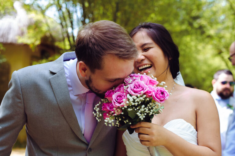 Groom smells the bride's pink rose bouquet while she laughs.