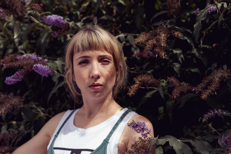 an outdoor headshot of a young woman standing next to a bush with purple flowers